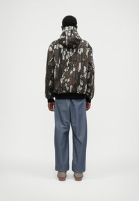 Braune und graue gemusterte Bomberjacke mit Kapuze und gerippten Bündchen, kombiniert mit locker sitzenden blauen Jeanshosen und grauen Stiefeln.