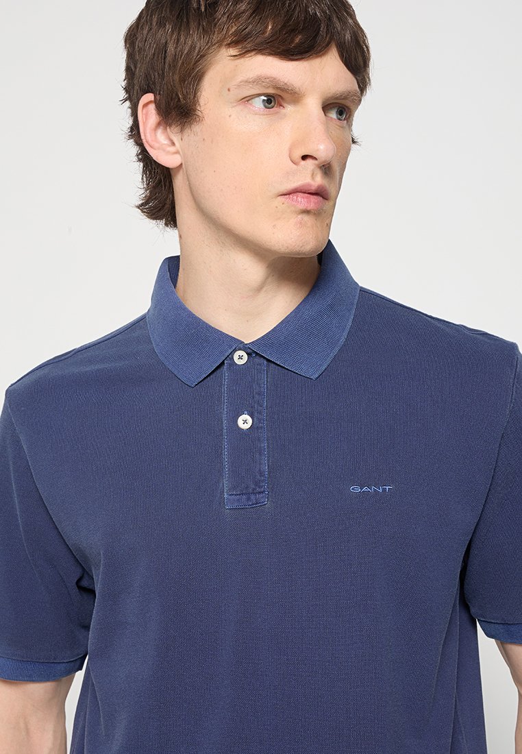 Młody mężczyzna z krótkimi brązowymi włosami, ubrany w granatową koszulę polo z dwoma białymi guzikami i małym logo na klatce piersiowej, spoglądający w bok.
