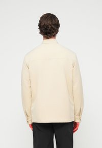 Μπεζ σακάκι με γιακά και μακριά μανίκια, ραφές στο εμπρός και πίσω μέρος, μανσέτες με κουμπιά και λεία υφή. Φορεμένο με μαύρο παντελόνι.