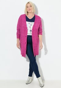 Cardigan lungo con apertura frontale in tessuto a maglia di colore rosa acceso, caratterizzato da chiusura con bottoni e polsini a coste, indossato sopra una T-shirt blu navy e jeans scuri.