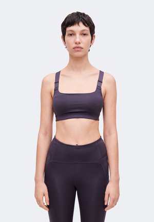 Vrouw met kort donker haar die een donkere sportbeha en bijpassende hoge taille legging draagt, staand met het gezicht naar voren tegen een effen achtergrond.
