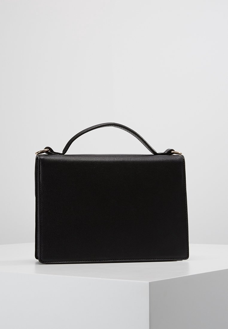 Sac à main rectangulaire en cuir noir avec une seule poignée supérieure, présenté sur un socle blanc sur fond gris.