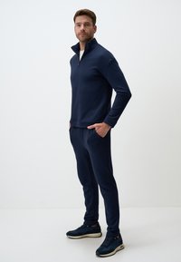 Sweat-shirt zippé bleu marine à manches longues, associé à un pantalon fuselé assorti. Présente une texture lisse et des poches latérales fonctionnelles.