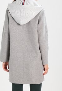 Person iført en lang, lys grå frakke med en grå hætte, der har det prægede ord "TOMMY" samt rød, hvid og blå kant.