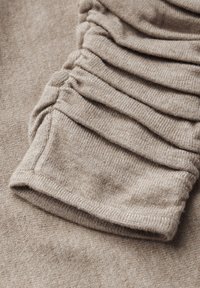 Tessuto lavorato a maglia beige con un design testurizzato e arricciato lungo l'orlo delle maniche. Dettaglio di polsino morbido e a coste. Superficie liscia con un leggero lucido.