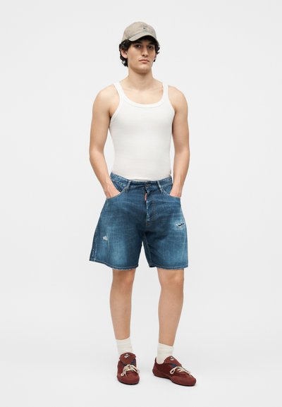 Νέος άνδρας που φοράει μπεζ καπέλο, λευκό αμάνικο πουκάμισο, μπλε τζιν σορτς, λευκές κάλτσες και κόκκινα αθλητικά παπούτσια, στέκεται με τα χέρια στις τσέπες.