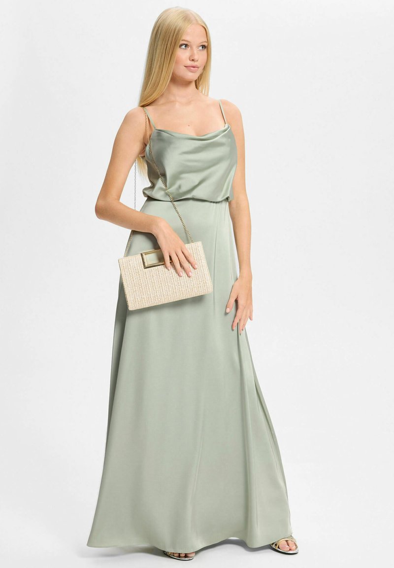 Jeune femme portant une robe sans manches en satin vert clair et tenant une pochette beige texturée avec une lanière en chaîne.