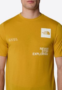 Hořčicově žluté bavlněné tričko s bílou nášivkou loga, geografickými souřadnicemi a textem "NIKDY NEPŘESTÁVEJ PROZKOUMÁVAT" v bílé barvě.