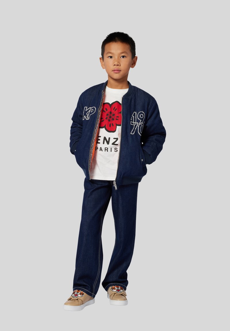 Veste bomber en denim avec des accents brodés, t-shirt blanc avec un graphique de fleur rouge, pantalon en denim foncé et baskets beiges avec un design de tigre.