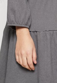 En avslappnad hand som vilar mot en grå, strukturerad långärmad klänning med elastiska ärmslut och samlad midja.