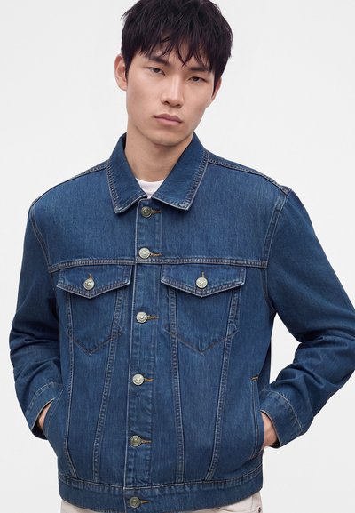 Jeune homme portant une veste en jean bleu foncé boutonnée, les mains dans les poches, debout devant un fond clair uni.
