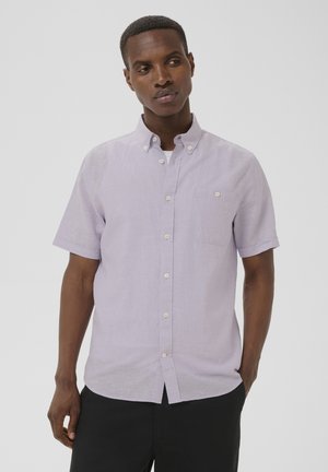 Homme portant une chemise à manches courtes violet clair par-dessus un sous-t-shirt blanc, debout avec une main dans la poche, regardant sur le côté.