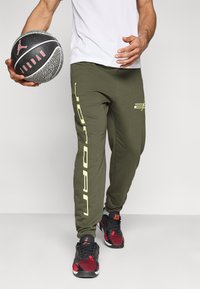 Ελιά πράσινη φόρμα με μεγάλο κείμενο «Jordan» σε ανοιχτό κίτρινο στο  πλάι, συνδυασμένη με μαύρα και κόκκινα αθλητικά παπούτσια και μια μπάλα μπάσκετ.