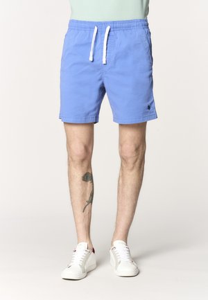 Uomo con pantaloncini azzurri chiaro con coulisse, sneakers bianche e un tatuaggio di piuma sul polpaccio sinistro, in piedi davanti a uno sfondo neutro.