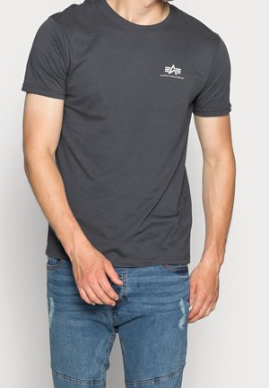 Homme portant un t-shirt gris foncé à manches courtes avec le logo blanc d'Alpha Industries sur la poitrine et un jean bleu délavé, debout devant un fond clair.