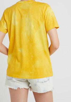 Osoba w żółtej koszulce z krótkim rękawem w stylu tie-dye oraz jasnoniebieskich, postrzępionych szortach jeansowych, widziana od tyłu.