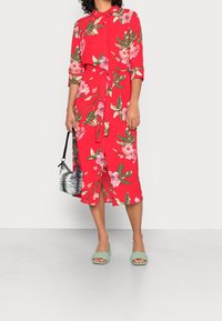 Robe florale rouge avec col, taille ceinturée et boutons, ornée de fleurs roses et de feuilles vertes. Associée à des sandales vertes à talon carré.