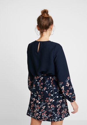 Kvinde med håret sat i en knold bærer en marineblå kjole med lange ærmer og et blomsterprintet skørt, vist bagfra mod en ensfarvet baggrund.