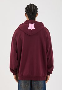 Personne avec des cheveux tressés portant un sweat à capuche ample maroon avec un design de papillon rose à l'arrière de la capuche et un jean bleu.