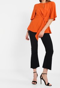 Blouse péplum orange avec un détail noué et un décolleté en trou de serrure, associée à un pantalon noir évasé à côtes et des talons noirs à brides.