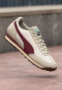 Beige athletic shoe with maroon side stripe, textured suede and leather upper, lace-up closure, rubber sole with molded studs.