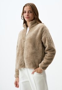 Beige, fluffy zip-up jack met hoge kraag en elastische manchetten, gemaakt van zacht synthetisch materiaal, met een relaxte pasvorm en voorzakken.