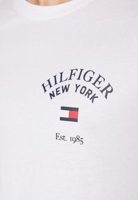 Camiseta de algodón blanca con la frase "HILFIGER NEW YORK" en texto negro, un logo con un patrón de bloques en rojo, blanco y azul, y "Est. 1985" debajo.