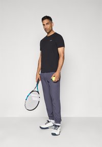 Zwarte T-shirt en grijze sportbroek, met een tennisracket en een gele bal. Schoenen zijn wit met blauwe accenten, en er staat een logo op het T-shirt.