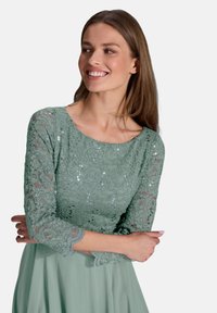 Robe en dentelle verte avec des sequins, décolleté rond, manches trois-quarts et une jupe fluide. Superposition en dentelle texturée avec des motifs floraux.