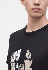 Νεαρό άτομο που φοράει μαύρο t-shirt με φθαρμένο λευκό σήμα ασπίδας και σχέδιο στεφάνου δάφνης στο στήθος σε μονόχρωμο φόντο.
