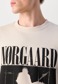 Beige sweatshirt with black "NORGAAARD" text, featuring a graphic print. Soft fabric, crew neck, minimal stitching details.