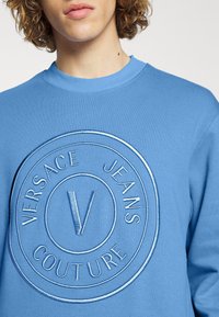 Jeune personne aux cheveux bouclés portant un sweat à capuche bleu avec un logo circulaire en relief portant l'inscription "Versace Jeans Couture."