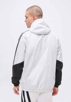 Homme de dos portant une veste à capuche blanche avec des manches noires et des bandes noires sur les bras, associée à un pantalon blanc avec des bandes noires.