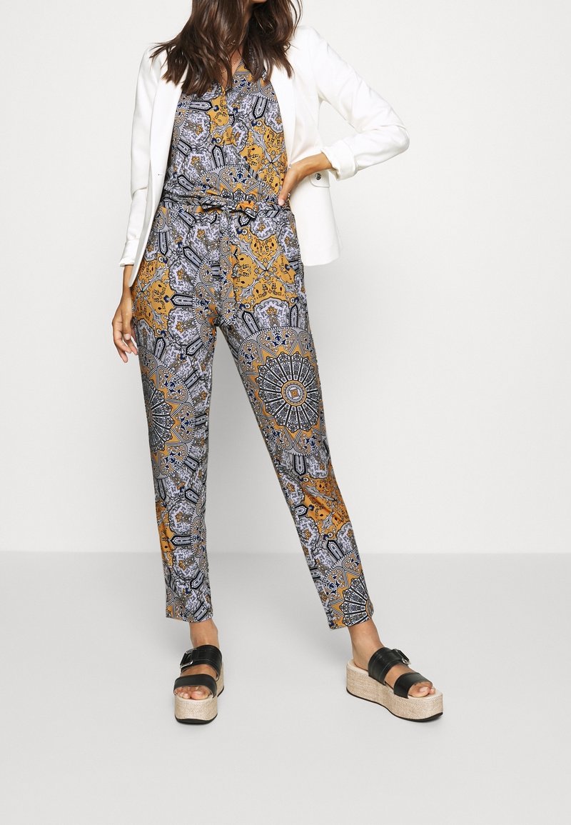Femme portant un pantalon taille haute à motifs orange, noir et gris, un blazer blanc et des sandales à plateforme noires, debout avec une main sur la hanche.