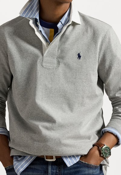 Grijze polo met lange mouwen en een kraag, met een marineblauw logo. Draag het over een blauw-wit gestreept shirt met een zichtbare polshorloge.