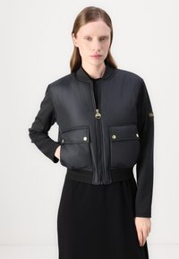 Μαύρο κοντό bomber jacket με φερμουάρ μπροστά, χρυσές λεπτομέρειες, δύο μπροστινές τσέπες και ριμπ μανσέτες. Φτιαγμένο από απαλό, επενδεδυμένο υλικό.