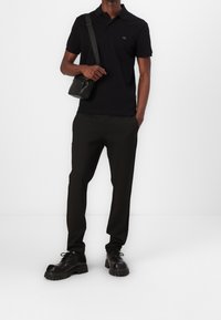 Polo noir avec un petit logo, associé à un pantalon noir à rayures et de grosses chaussures noires. Les accessoires comprennent un petit sac banane noir.