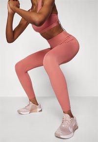 Růžové sportovní legíny s vyšším pasem, hladkým povrchem a švy, doplněné o světle růžové síťované tenisky s bílým logem.