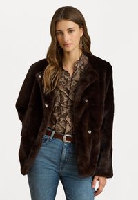 Barna műszőrme kabát, dupla soros gombolással és ezüst gombokkal, fodros mintás blúzzal és kék farmernadrággal párosítva.