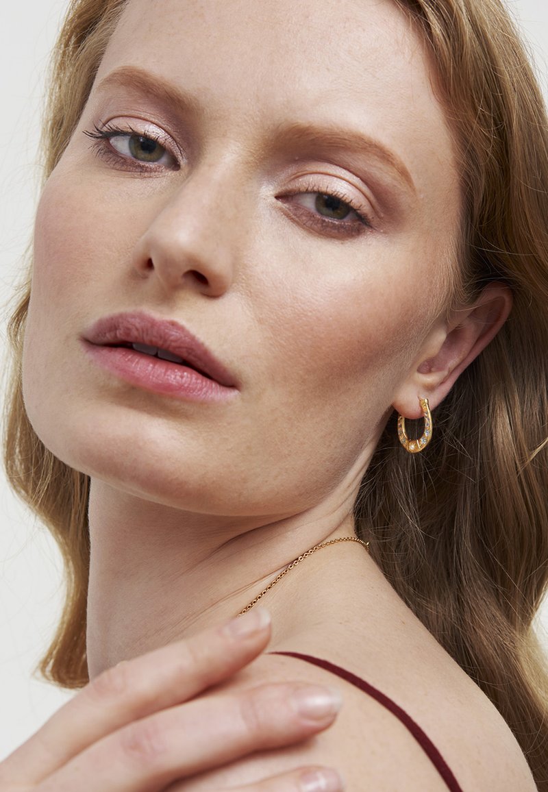 Zlatá kruhová náušnica s textúrovaným vzorom. Záber na tvár osoby s jemným líčením, hladkou pleťou a vlnitými, po plecia dlhými vlasmi.