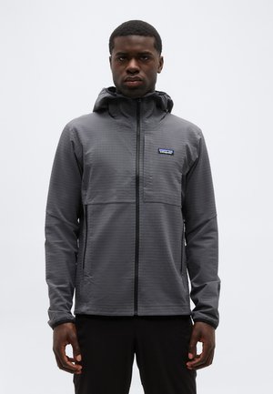 Jeune homme portant une veste à capuche gris à fermeture éclair avec un logo Patagonia sur la poitrine, debout devant un fond blanc uni.