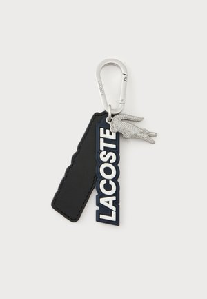 Metalowy brelok z karabińczykiem z czarną metką, pionową metką z logo "LACOSTE" oraz małym srebrnym wisiorkiem w kształcie krokodyla na białym tle.