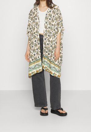 Femme portant un kimono blanc à motif floral sur un crop top blanc, un jean noir large et des sandales à plateforme noires.