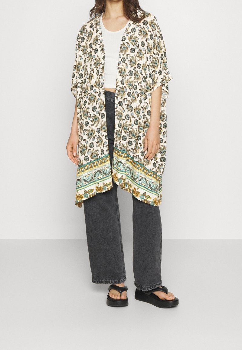 Femme portant un kimono blanc à motif floral sur un crop top blanc, un jean noir large et des sandales à plateforme noires.