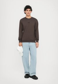 Felpa marrone scuro a maniche lunghe con colletto a girocollo, caratterizzata da un logo nero sul petto, abbinata a jeans a gamba larga blu chiaro e un cappellino bianco.