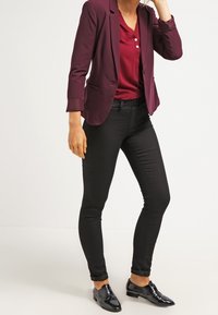 Blazer burdeos sobre una camisa roja abotonada, combinado con jeans pitillo negros y zapatos negros brillantes. La tela parece suave y con un ajuste entallado.