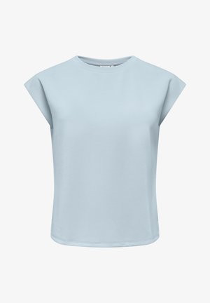 Femme debout portant un haut bleu clair à manches courtes, un jean bleu et des chaussures blanches à bouts pointus sur fond blanc.
