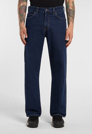 Mies, jolla on tummansiniset suorat farkut ja mustat lenkkarit, esiintyen tatuoidut kyynärvarret ja kädet rentoina sivuilla.