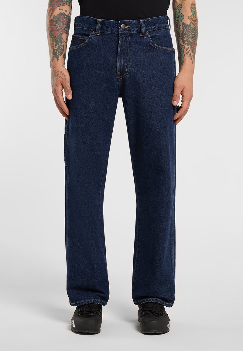 Mand iført mørkeblå jeans med lige ben og sorte sneakers, der viser tatoverede underarme, mens hænderne hviler afslappet ved siderne.