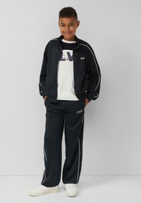 Zwarte trainingsjack met witte biezen, gedragen met bijpassende broeken, met een logo. Daaronder een wit T-shirt met kleurrijke tekst. Witte sneakers.